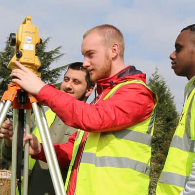 two students and a teacher measure with some surveying equipment outside on campus