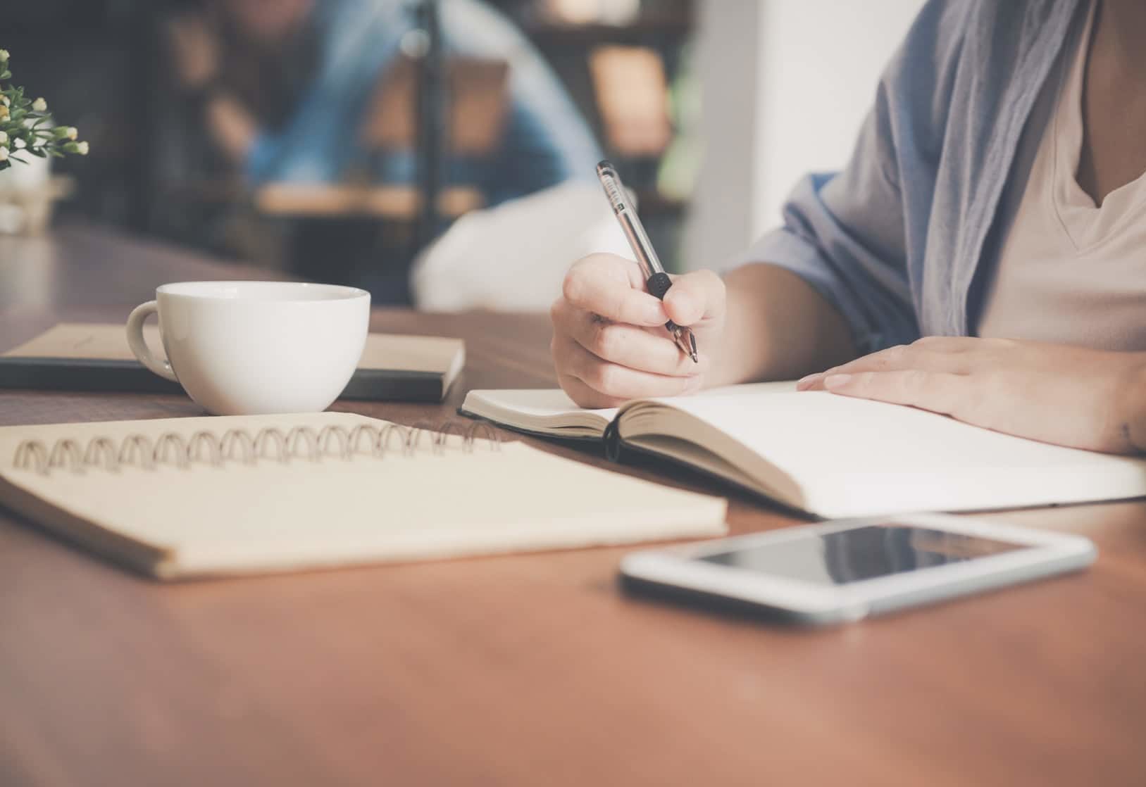 Student writing notes at desk with cup and smartphone.