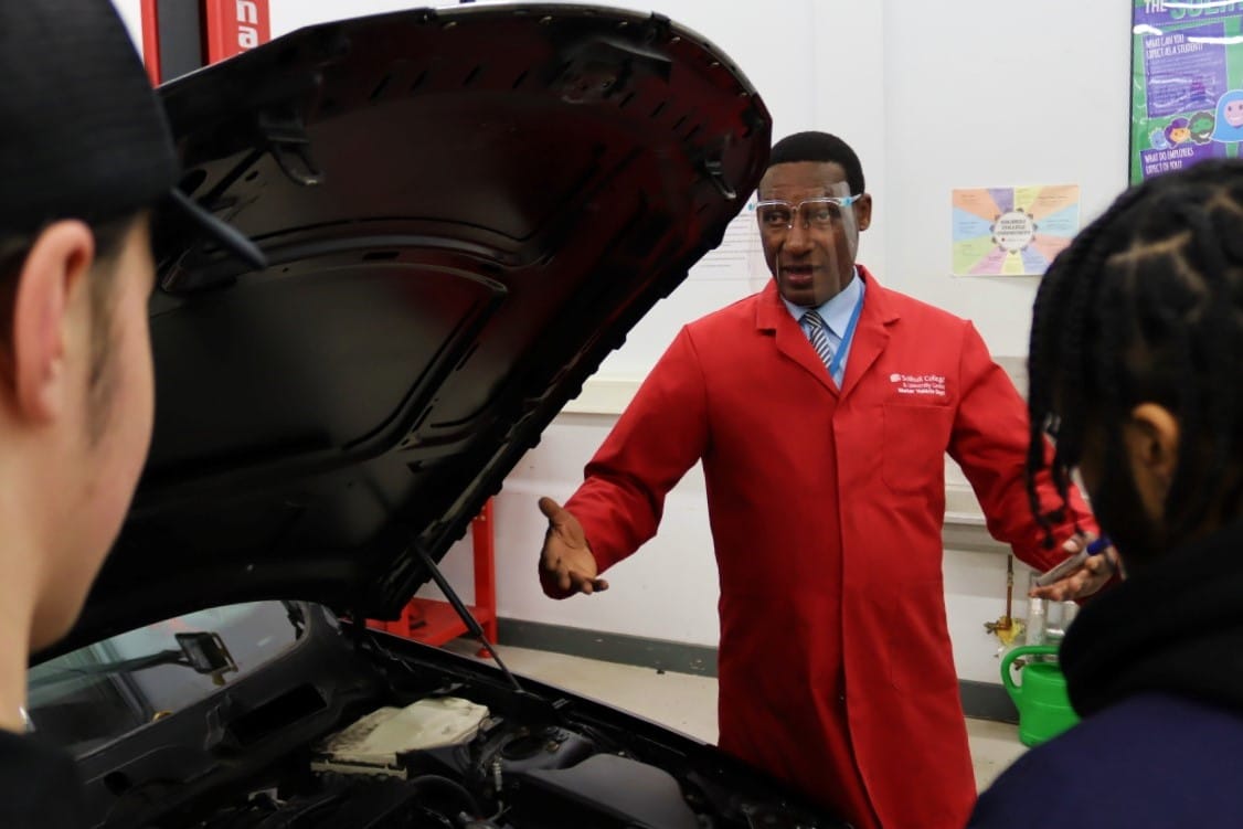Motor Vehicle Lecturer in red coat explaining car to students with hood of car open between them