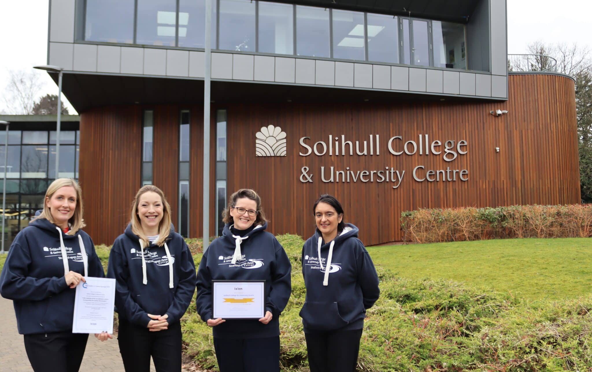 4 staff members standing in front of college building holding certificates