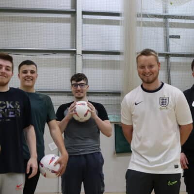 5 students standing in sports hall, three holding a football