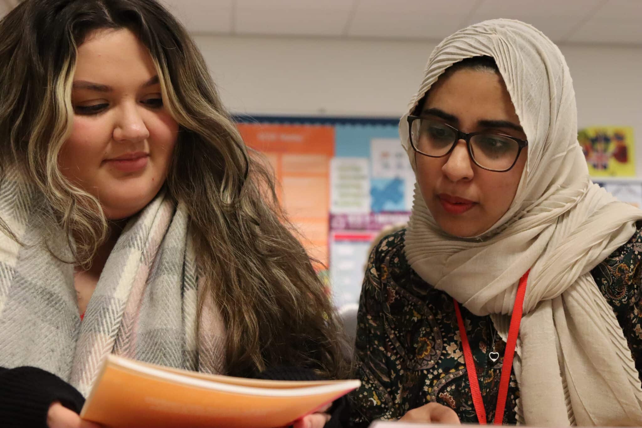 Two students in a classroom looking at a book cover.