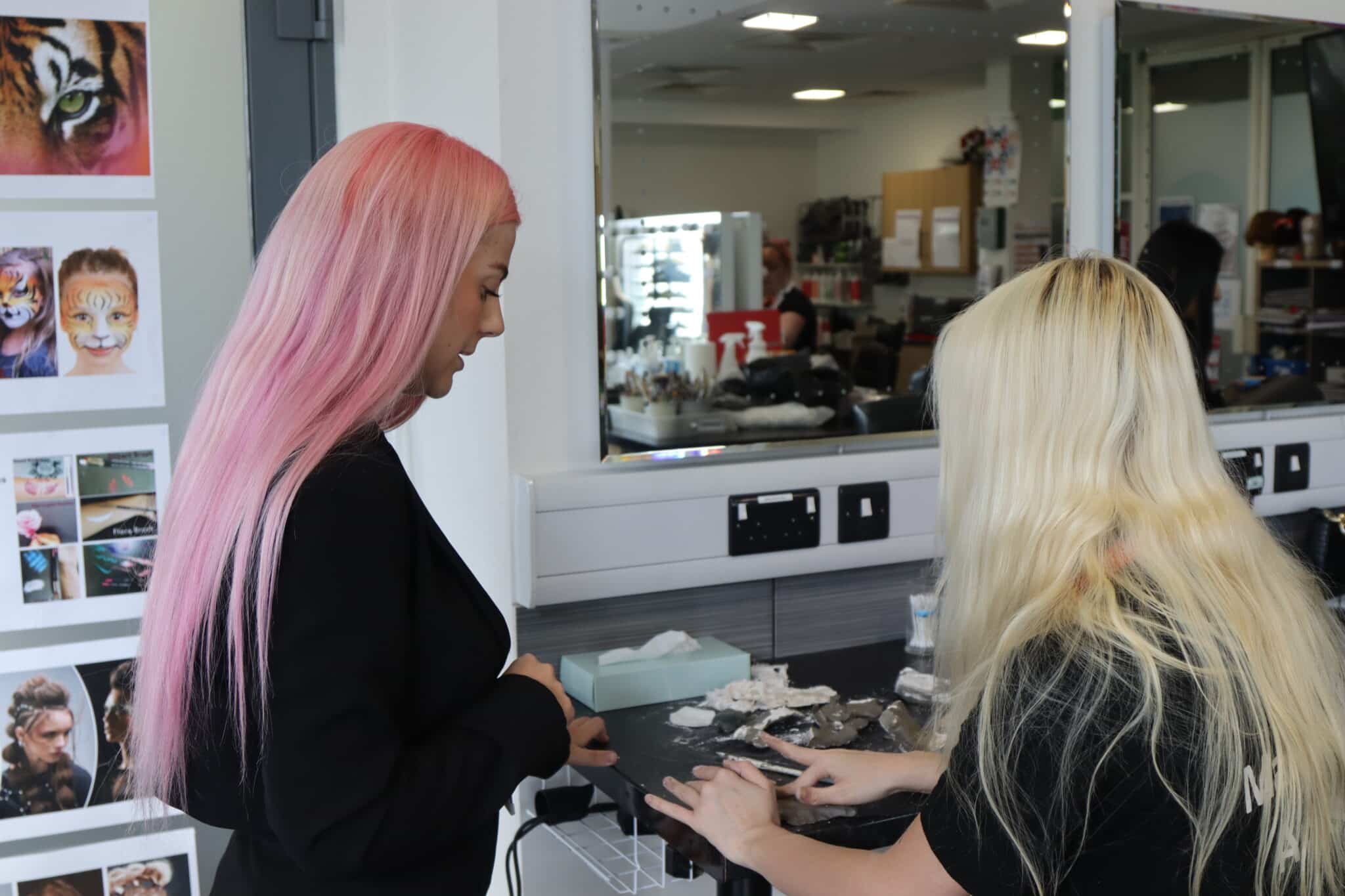 A teacher with pink hair and student with blonde hair sat at a make-up station